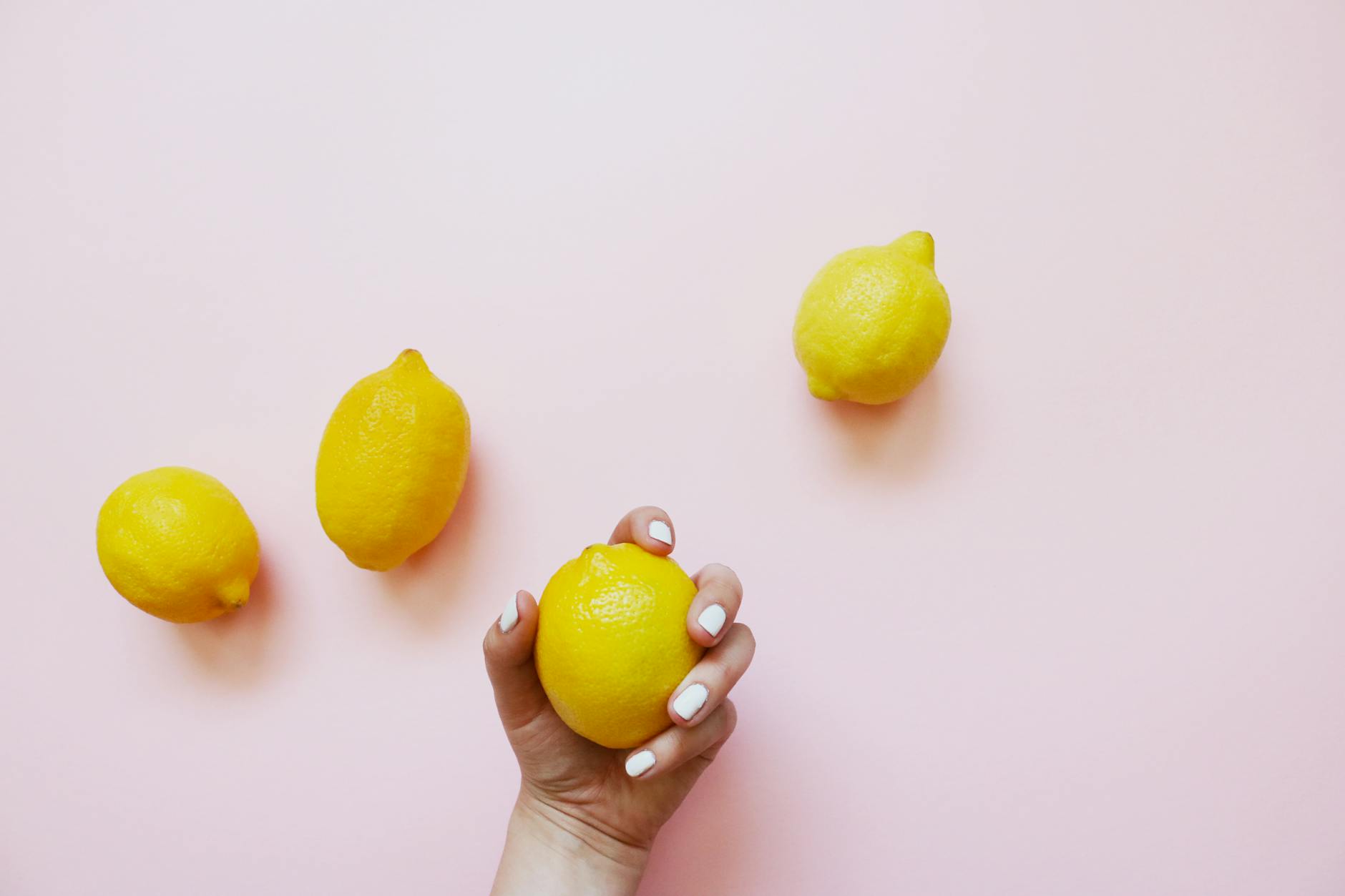 Una mano con unghie bianche che tiene un limone su uno sfondo rosa morbido, circondato da altri limoni.