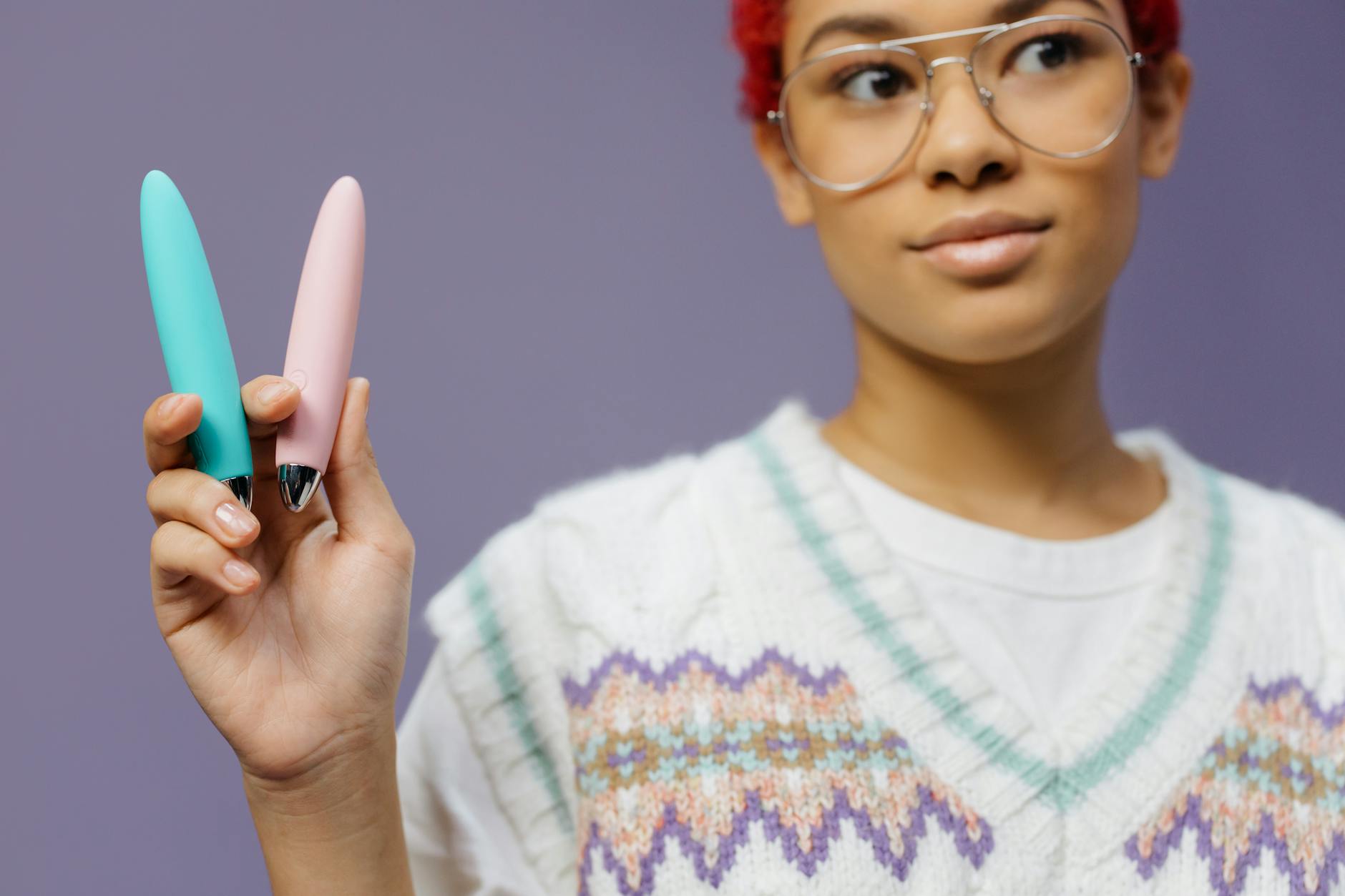 Una donna con gli occhiali tiene in mano due vibratori colorati contro uno sfondo viola.