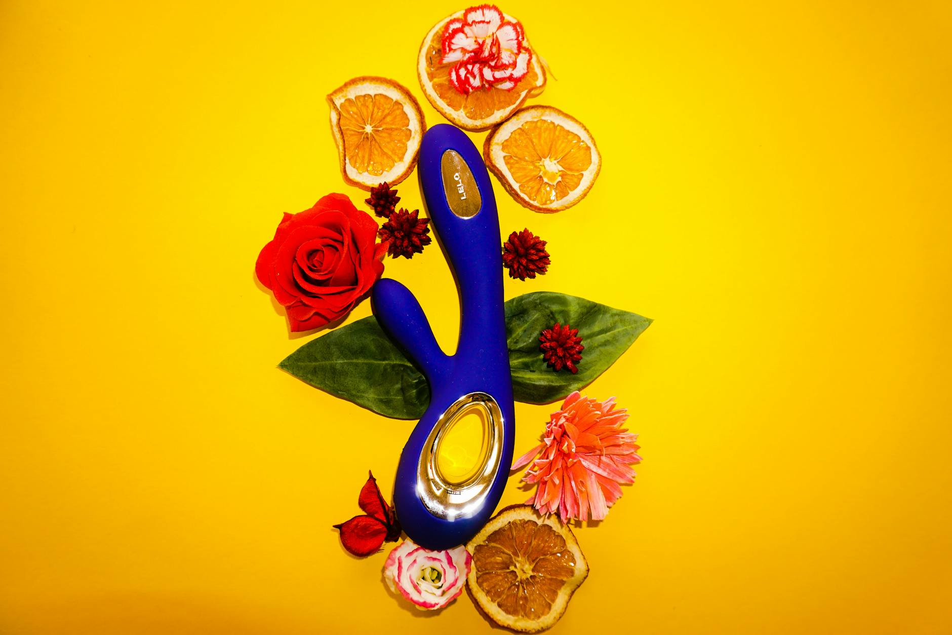 Vibratore blu circondato da fiori e fette d'arancia su sfondo giallo, simbolo di piacere naturale e fresco